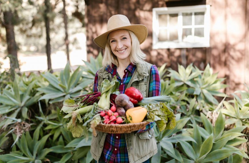 woman in a hat holding a basket of assorted vegetables in a backyard garden