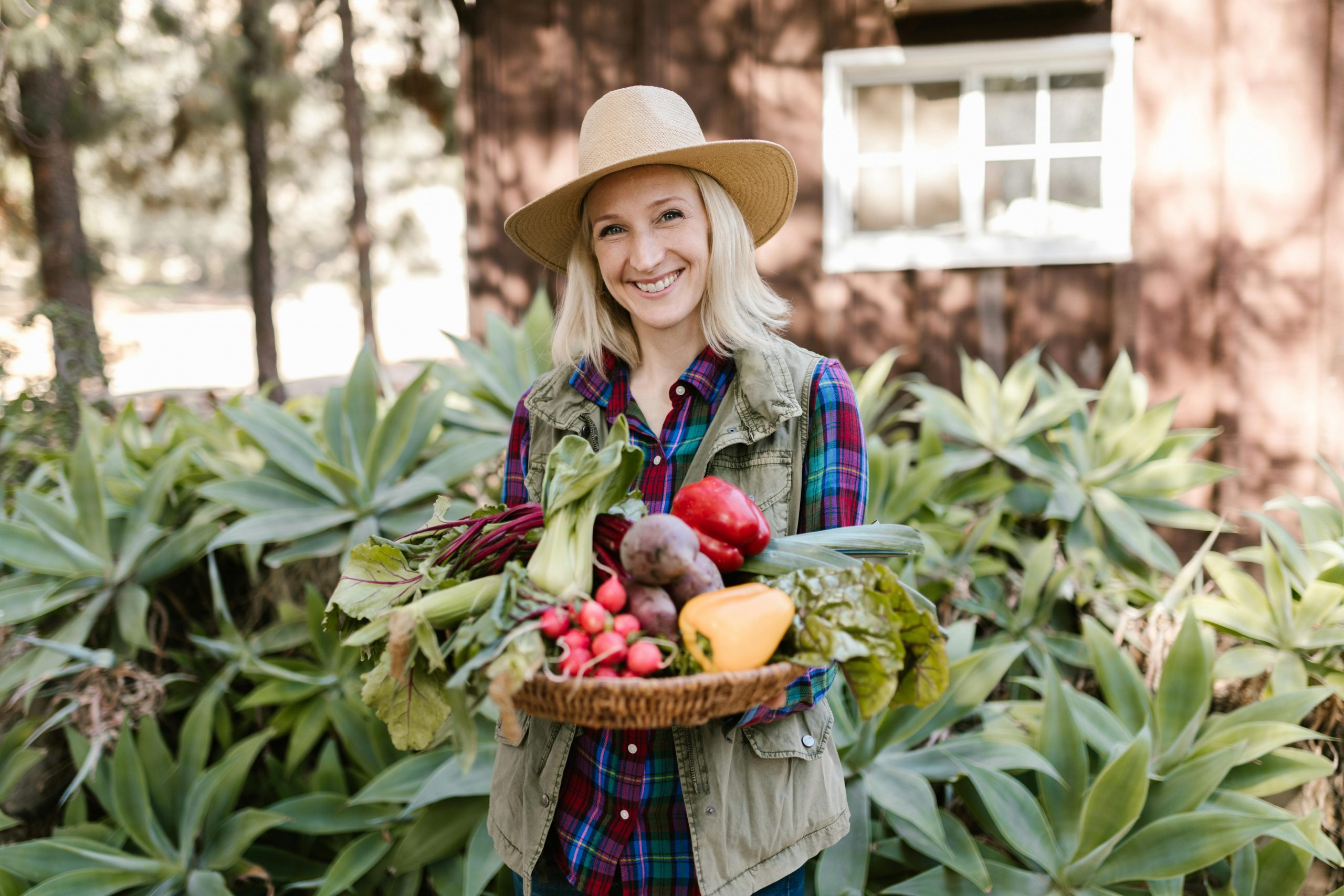 woman in a hat holding a basket of assorted vegetables in a backyard garden